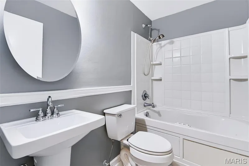 Pedestal sink with chrome faucet, circular wall mirror, toilet, and a combination bathtub/shower with a tiled surround — 1314 Warrior Song Way, O'Fallon, MO 63366