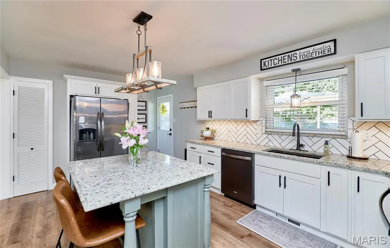 Spacious kitchen featuring an island with stone countertop, white shaker cabinetry with black hardware, stainless steel appliances, wood-finish flooring, and a decorative tile backsplash — 415 Westridge Drive, O'Fallon, MO 63366