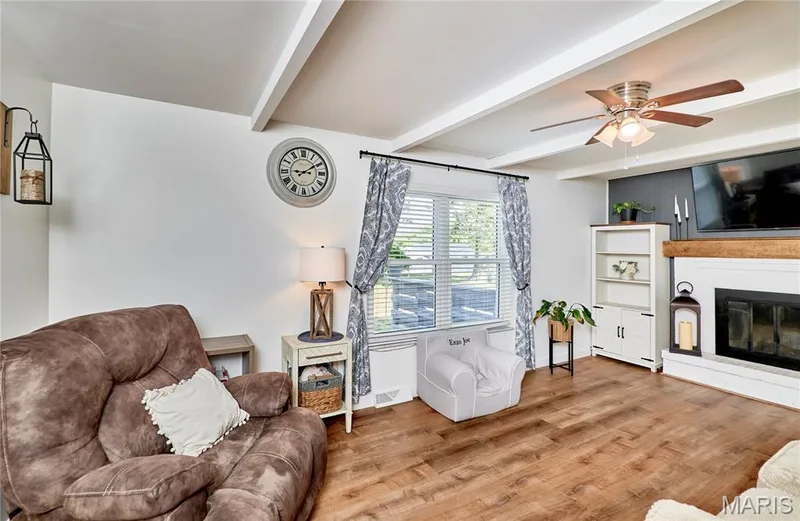 Vaulted ceiling with exposed beams, wood-finish flooring, white fireplace with a wood mantel, ceiling fan with integrated lighting, and built-in shelving — 415 Westridge Drive, O'Fallon, MO 63366