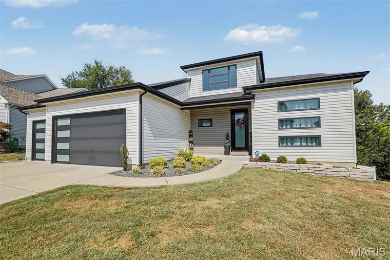 Contemporary exterior design with light-colored siding, dark trim, a two-car garage with frosted glass panels, an arched concrete walkway, and a stacked stone retaining wall — 18 MacArthur Court, St Charles, MO 63304