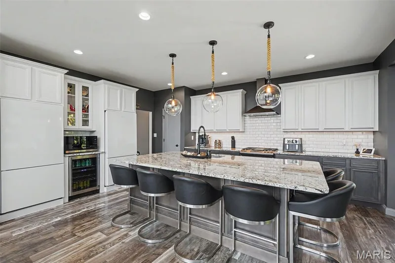 Kitchen featuring a large island with stone countertop, white upper cabinetry, dark lower cabinetry, double refrigerators, wine cooler, pendant lights, and wood-finish flooring — 18 MacArthur Court, St Charles, MO 63304