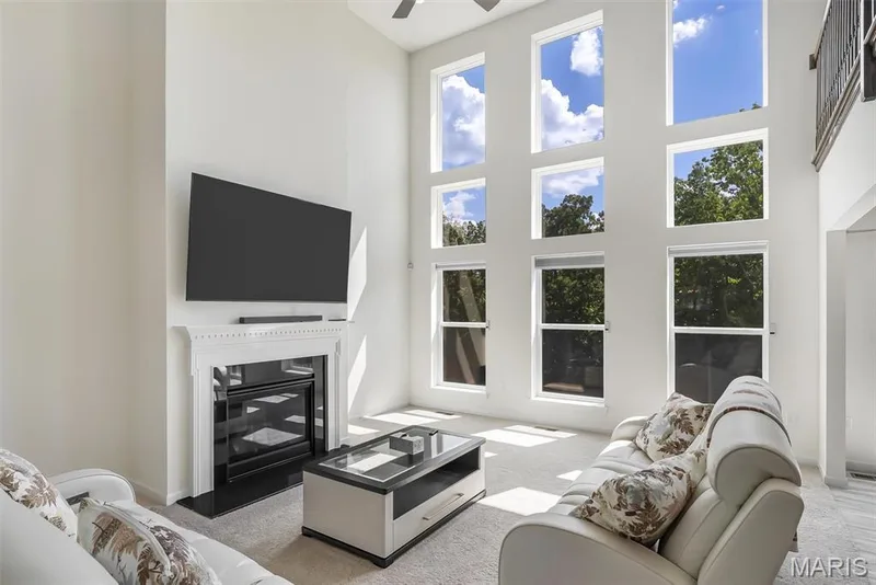 Two-story living area with a fireplace featuring a white mantel and black hearth, wall-mounted television, and a ceiling fan — 337 White Orchid Court, Wentzville, MO 63385