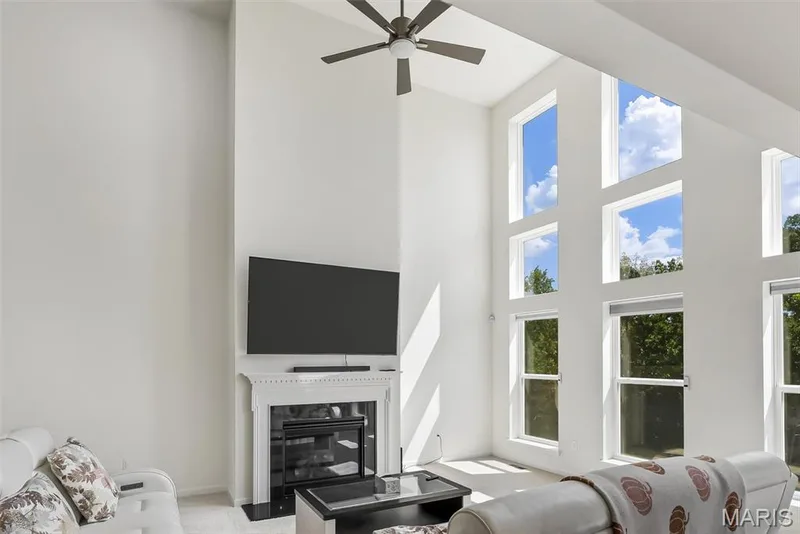Living space featuring a tall ceiling, a ceiling fan with light, a white mantel, and multiple large windows — 337 White Orchid Court, Wentzville, MO 63385