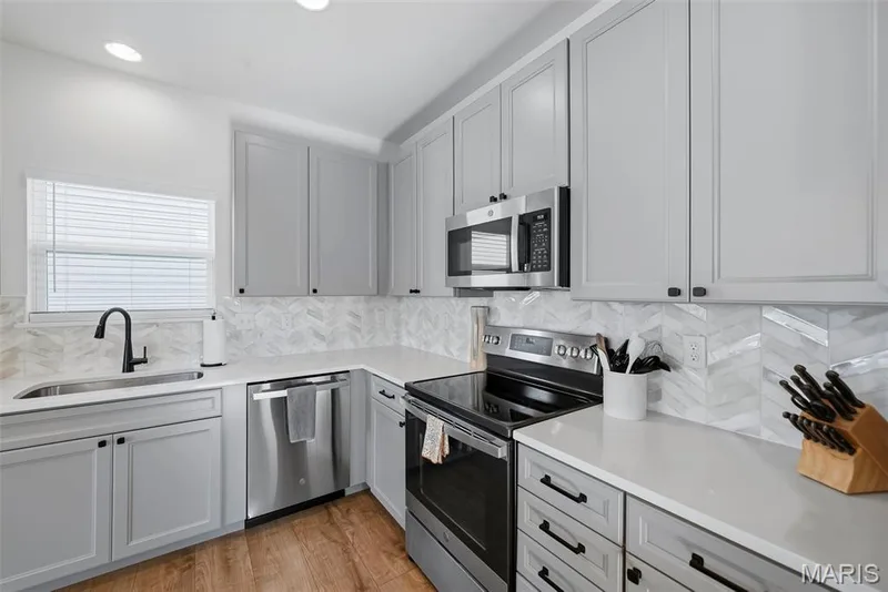 Kitchen featuring gray shaker-style cabinetry, stainless steel appliances, a chevron pattern tile backsplash, light-toned countertops, and wood-finish flooring — 191 Crestfield Court, St Charles, MO 63301