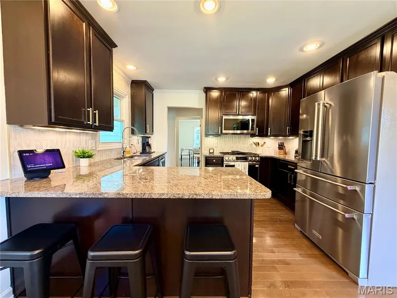Kitchen featuring dark wood cabinetry, light-toned granite countertops, a textured tile backsplash, stainless steel appliances, and recessed lighting — 584 Sunbridge Drive, Chesterfield, MO 63017