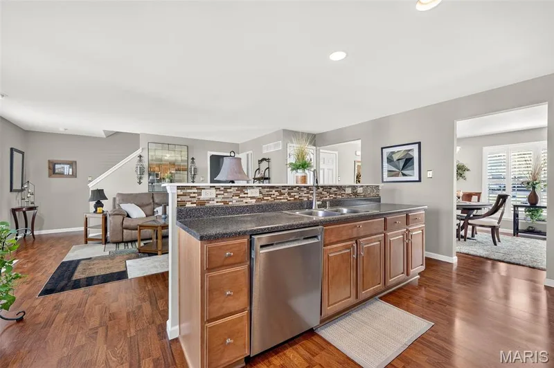 Wood-finish flooring throughout, kitchen island with built-in stainless steel dishwasher and dual basin sink, wood cabinetry, stone-look countertop, and a tiled backsplash — 29 Edenshire Court #69B, Dardenne Prairie, MO 63368