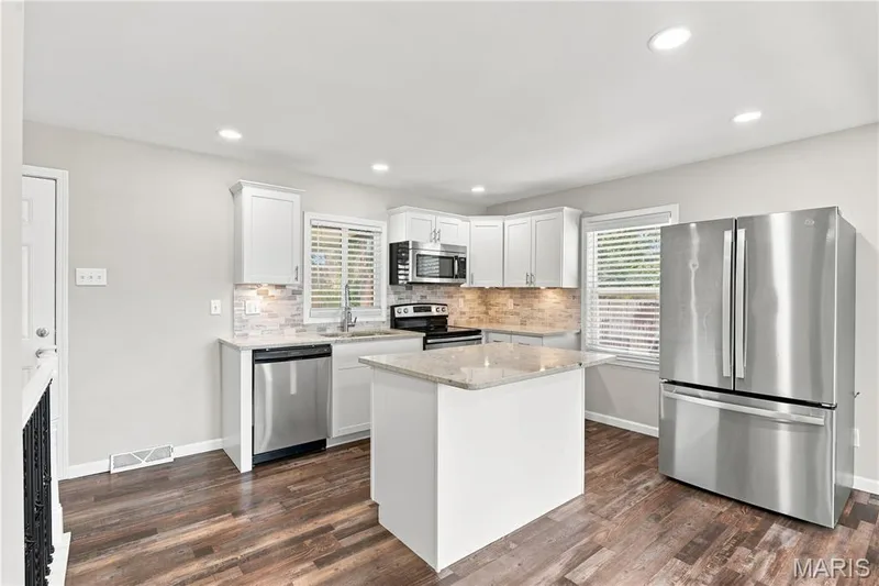 Modern kitchen featuring stainless steel appliances, white cabinetry with soft-close doors, quartz countertops, a large center island with seating space, subway tile backsplash, recessed lighting, wood-finish flooring, and plantation shutters — 1620 Dale Avenue, St Charles, MO 63301
