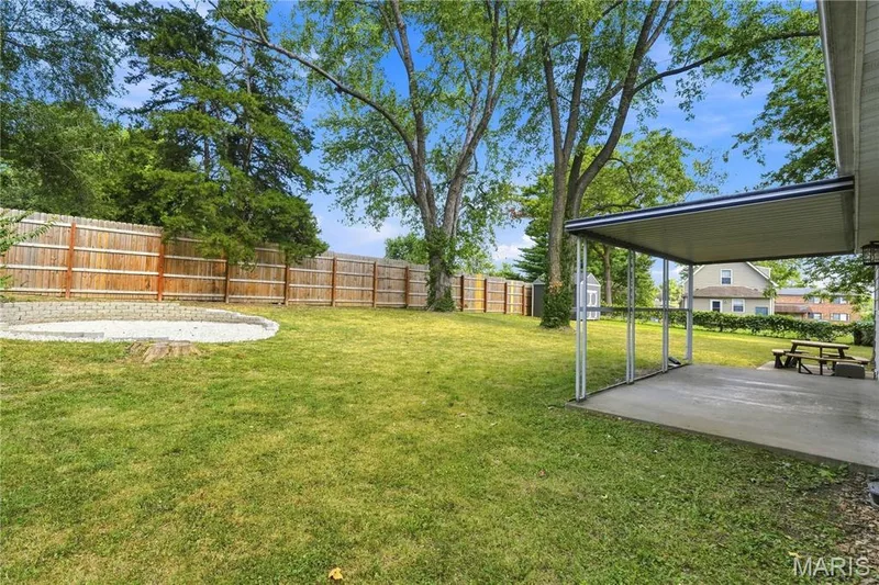 Expansive backyard featuring a manicured lawn, wood privacy fencing, a covered concrete patio, mature trees, and a gravel fire pit area — 1620 Dale Avenue, St Charles, MO 63301