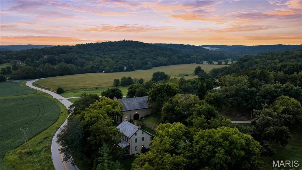 Extensive tree-lined acreage with a private winding road, featuring a stone-clad residence and a separate barn structure, bordered by open fields and distant hills — 2275 Highway F, Defiance, MO 63341