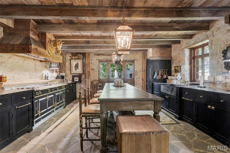 Kitchen with exposed wood beam ceiling, flagstone flooring, stone walls, black cabinetry with gold-tone hardware, and a large central island — 2275 Highway F, Defiance, MO 63341
