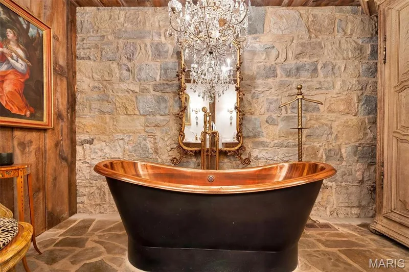 Freestanding copper bathtub with a dark exterior, stone wall, wood paneling, flagstone flooring, and an ornate chandelier — 2275 Highway F, Defiance, MO 63341