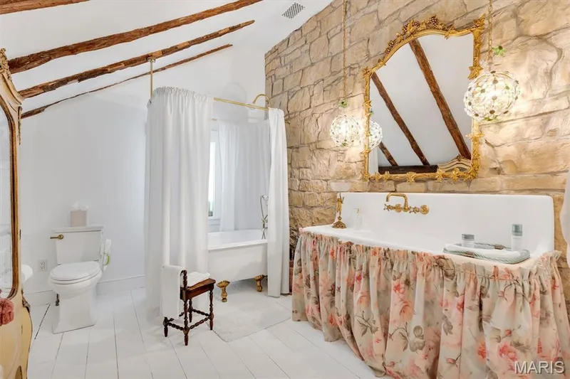 Exposed wood beam ceiling, stone accent wall, decorative gold-framed mirror, farmhouse-style sink with brass fixtures, and a clawfoot tub with shower enclosure — 2275 Highway F, Defiance, MO 63341