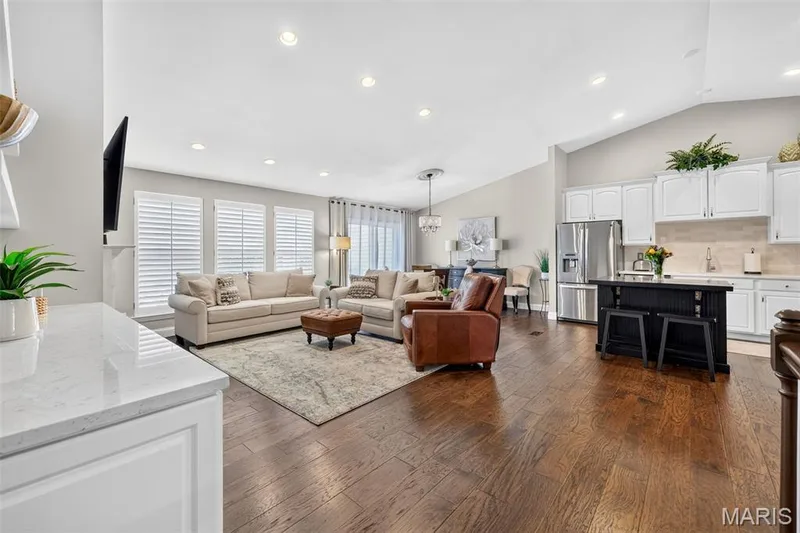 Expansive main living area featuring dark wood-finish flooring, white plantation shutters, vaulted ceilings, recessed lighting, and a kitchen island with seating — 14737 Ladue Bluffs Crossing Drive, Chesterfield, MO 63017