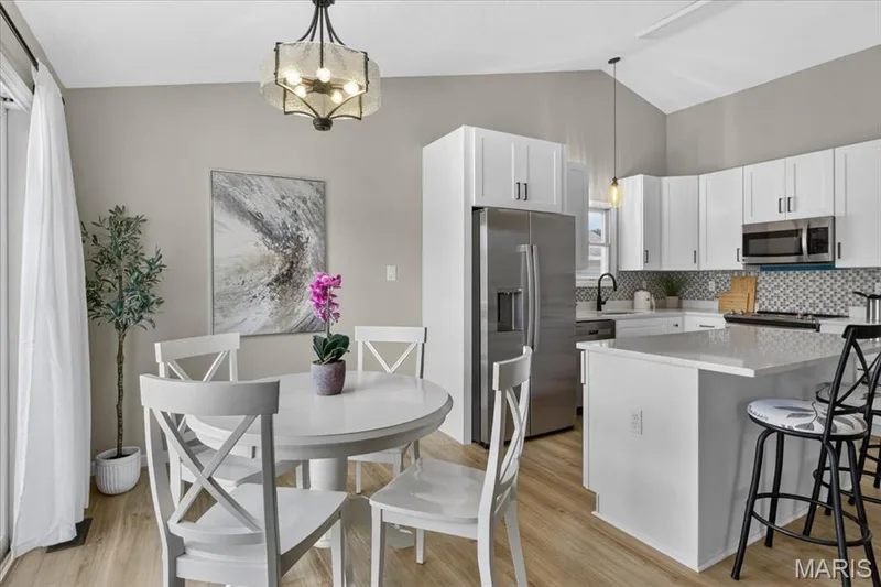 Vaulted ceiling with decorative light fixture, white cabinetry with black hardware, stainless steel appliances, mosaic tile backsplash, and light wood-finish flooring — 89 Gregory Court, Wentzville, MO 63385
