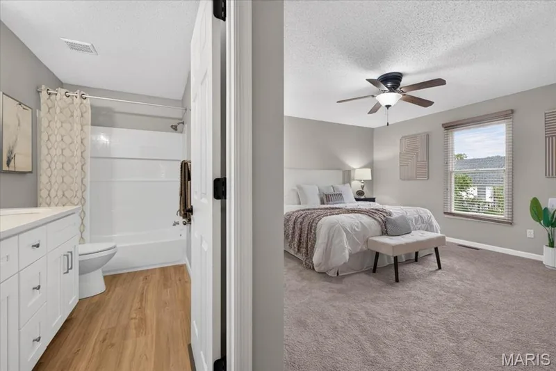 Recessed shower/tub combination, white vanity with integrated sink, wood-finish flooring, overhead ceiling fan with lighting, and a large window with blinds — 89 Gregory Court, Wentzville, MO 63385