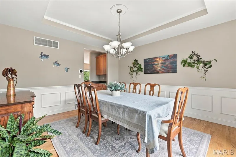 Formal dining with tray ceiling with central chandelier, wood floors, wainscoting, and neutral wall tones — 1000 Gardenbridge Place, St Charles, MO 63303