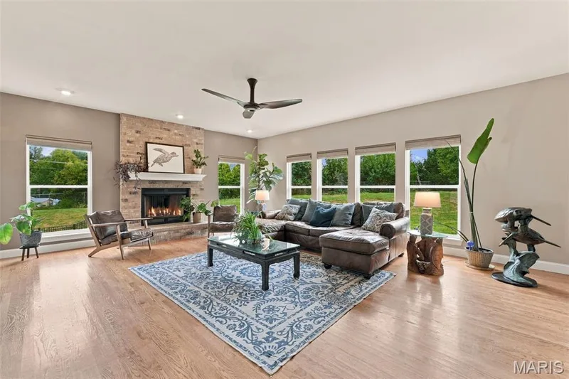 Spacious living area featuring wood flooring, automated roman shades, a brick-surround fireplace and contemporary ceiling fan — 1000 Gardenbridge Place, St Charles, MO 63303