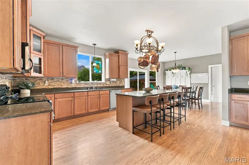 Open-concept kitchen featuring quartz countertops, central island with seating, a pot rack chandelier, ample wood cabinetry, and tile backsplash — 1000 Gardenbridge Place, St Charles, MO 63303