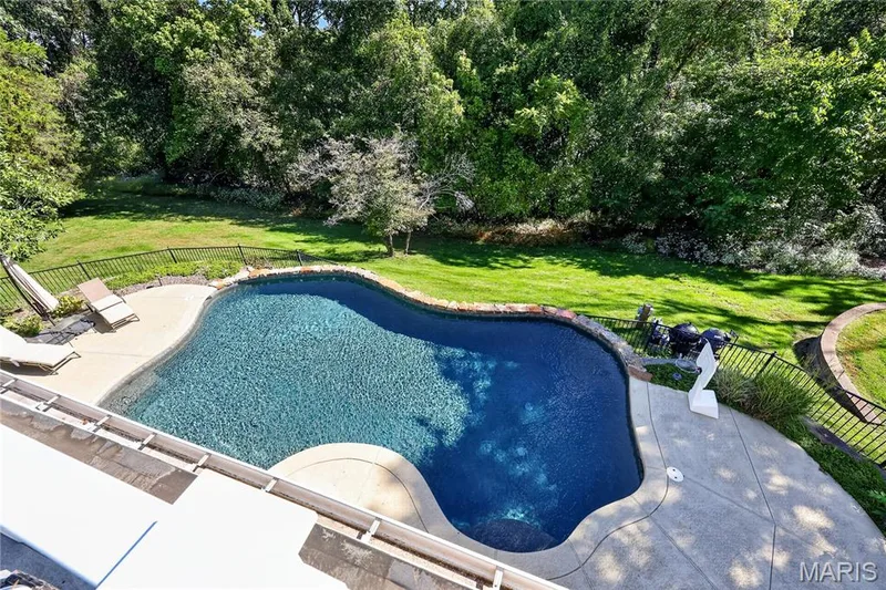 Freeform swimming pool with a surrounding concrete patio, black metal fencing, and abundant mature trees — 17711 Birch Leaf Court, Chesterfield, MO 63005