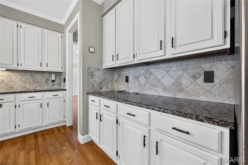 White cabinetry with black hardware, stone tile backsplash, dark stone countertops, wood-finish flooring, and crown molding — 17711 Birch Leaf Court, Chesterfield, MO 63005