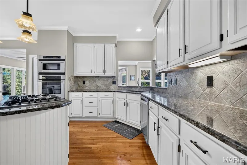 Kitchen featuring wood-finish flooring, white cabinetry, integrated stainless steel appliances, dark stone countertops, and a tiled backsplash — 17711 Birch Leaf Court, Chesterfield, MO 63005