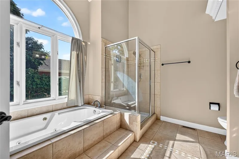Soaking tub with tiled surround, glass-enclosed shower, arched window, and neutral wall tones — 17711 Birch Leaf Court, Chesterfield, MO 63005