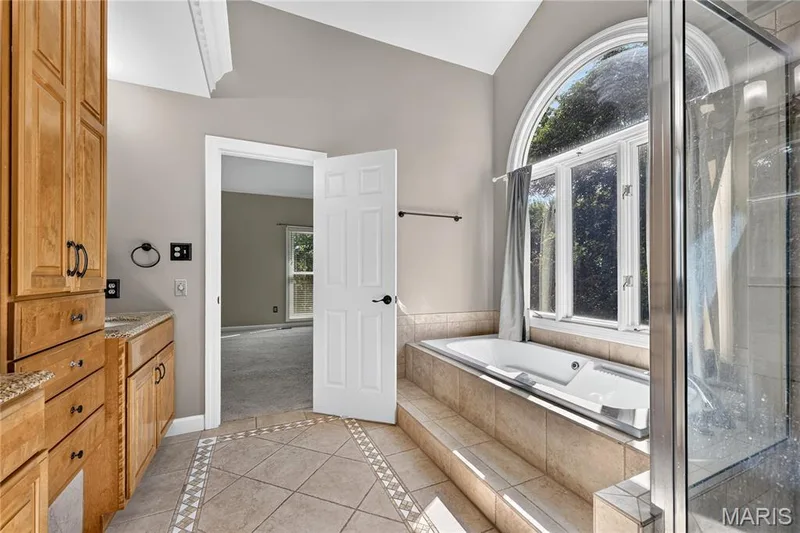 Jetted soaking tub with tiled surround, arched window, glass-enclosed shower, wood cabinetry, and tiled flooring — 17711 Birch Leaf Court, Chesterfield, MO 63005