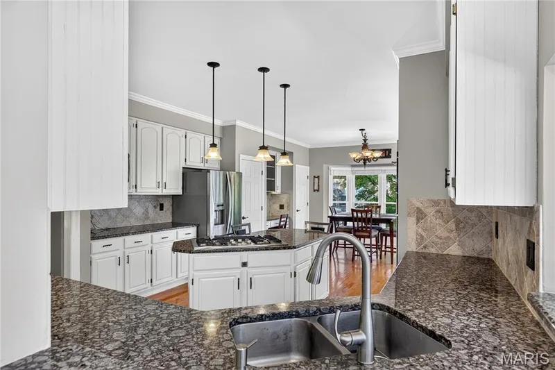 Spacious kitchen featuring white cabinetry, dark speckled countertops, and a stainless steel double basin sink with a gooseneck faucet — 17711 Birch Leaf Court, Chesterfield, MO 63005