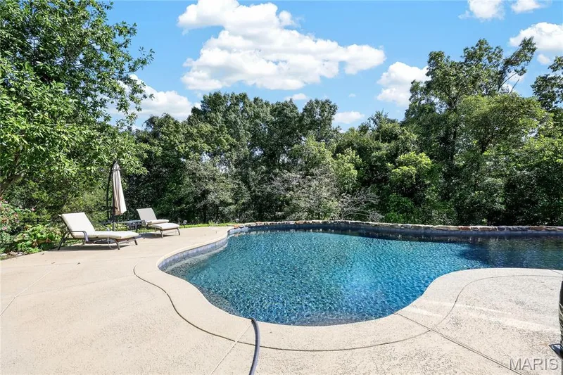 Freeform swimming pool with a stone coping, surrounded by a textured concrete deck and mature trees — 17711 Birch Leaf Court, Chesterfield, MO 63005