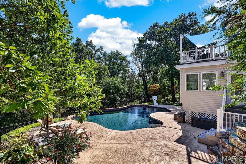 Freeform swimming pool with stamped concrete decking, surrounded by mature trees, an upper-level deck, and light-colored siding — 17711 Birch Leaf Court, Chesterfield, MO 63005