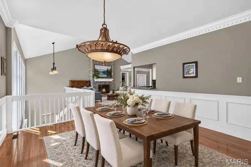Dining area featuring wood-finish flooring, white wainscoting, crown molding, and an ornate metal chandelier, with a stone fireplace surround in the background — 17711 Birch Leaf Court, Chesterfield, MO 63005