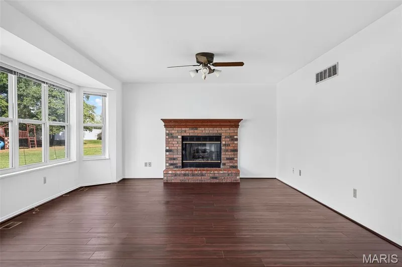 Spacious room featuring wood-finish flooring, a prominent brick fireplace with a wood mantel, a multi-pane bay window, and a ceiling fan — 39 Green Pines Circle, St Peters, MO 63376