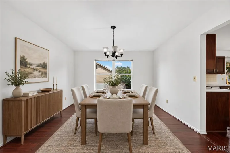 Formal dining area featuring rich wood-finish flooring, a contemporary chandelier, a large window, and an opening to a kitchen area with wood cabinetry and light countertops — 39 Green Pines Circle, St Peters, MO 63376