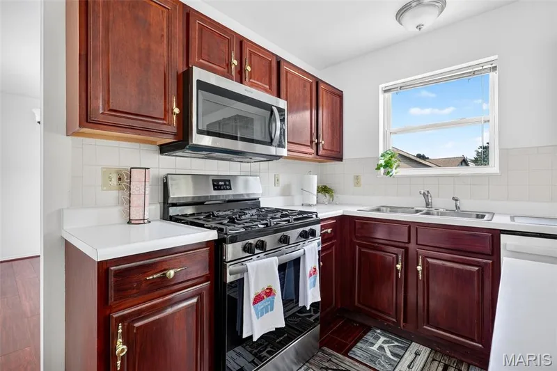 Compact kitchen featuring rich wood-finish cabinetry with brass hardware, stainless steel gas range and microwave, white quartz countertops, and a window allowing natural light — 39 Green Pines Circle, St Peters, MO 63376
