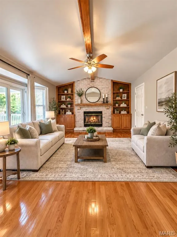 Hardwood flooring, a brick fireplace with wood mantel, built-in wood shelving units, a vaulted ceiling, and a ceiling fan, virtually staged — 3 Fairfax Drive, St Peters, MO 63376