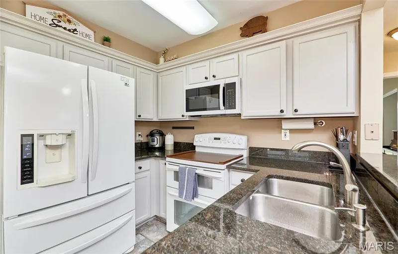 White cabinetry, dark stone countertops, stainless steel double basin sink, white built-in microwave, and integrated refrigerator — 16500 Victoria Crossing Drive, Wildwood, MO 63040