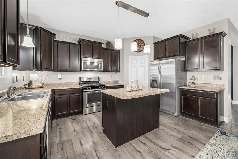 Kitchen featuring wood-finish flooring, dark wood cabinetry, light speckled countertops, stainless steel appliances, and a central island — 543 Country Chase Drive, Lake St. Louis, MO 63367
