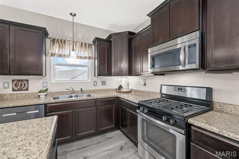 Dark wood cabinetry, stainless steel appliances, speckled countertops, stainless steel sink, and wood-finish flooring — 543 Country Chase Drive, Lake St. Louis, MO 63367