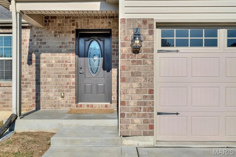 Brick exterior with a decorative front door, concrete steps, a paneled garage door, and a wall-mounted exterior light fixture — 56 Huntleigh Park Court, Wentzville, MO 63385
