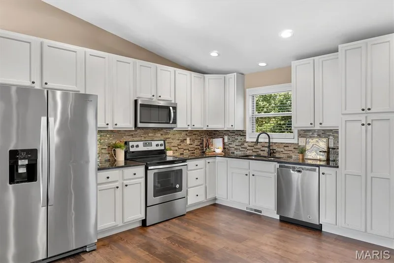 Kitchen featuring white cabinetry, dark countertops, stainless steel appliances, a tiled backsplash, and wood-finish flooring — 56 Huntleigh Park Court, Wentzville, MO 63385