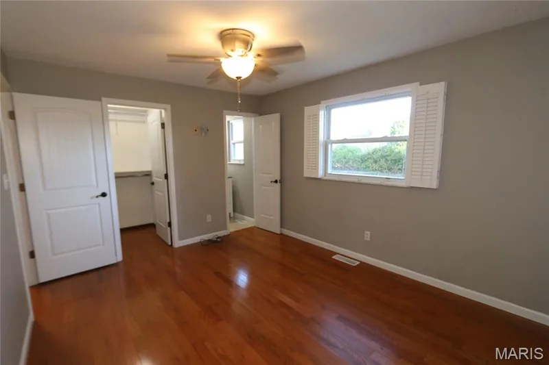 View from inside corner of Primary showing walk-in closet & bath entrances with shutters to the side. Wood-finish flooring, ceiling fan with integrated lighting, white interior doors, window with white louvered shutters, and a built-in closet with shelving — 3 Oxbow Road, St Peters, MO 63376