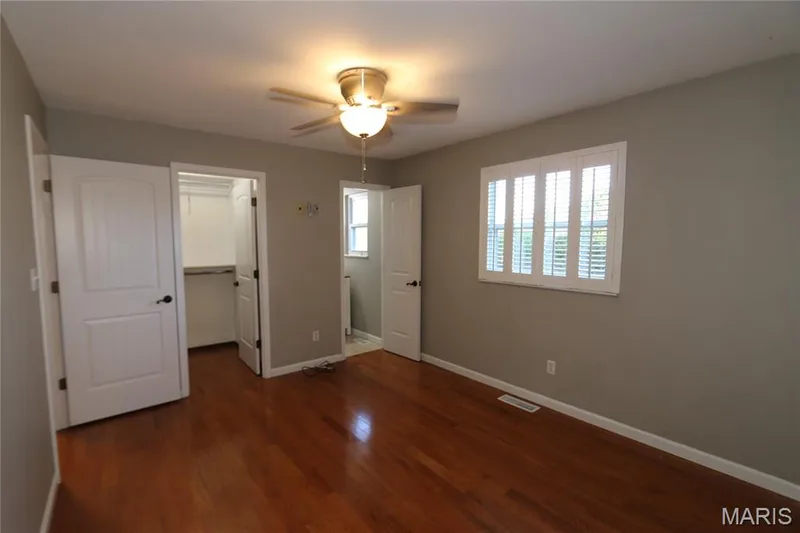 View from inside corner showing walk-in closet & bath with shutters closed with slats open. Warm wood-finish flooring, neutral wall tones, and a ceiling fan with integrated lighting — 3 Oxbow Road, St Peters, MO 63376
