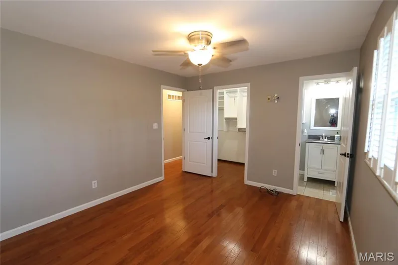 View from back corner of primary showing entrance from hall, walk-in bath & primary bath. Hardwood flooring, ceiling fan, neutral wall tones, built-in closet organization, and a vanity with a rectangular mirror — 3 Oxbow Road, St Peters, MO 63376