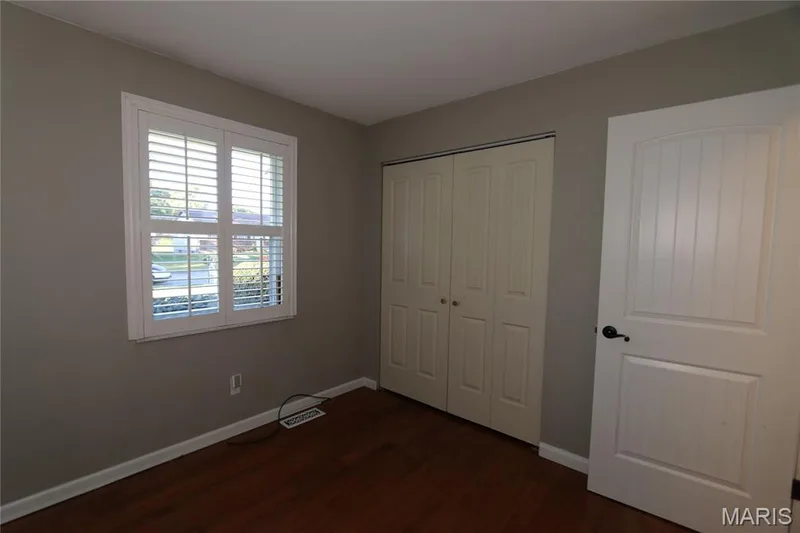 #3 Bedroom - view from flowered corner with shutters closed yet slats open. Wood-finish flooring with white baseboards, a window with white plantation shutters, and a white interior door with dark hardware — 3 Oxbow Road, St Peters, MO 63376