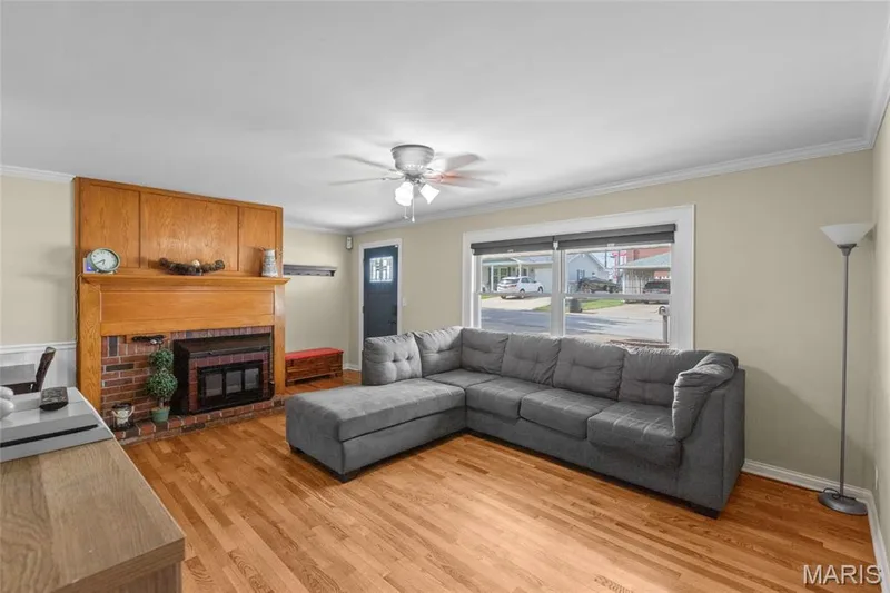 Wood-finish flooring throughout, brick fireplace with a wood mantel, crown molding, ceiling fan, and a large window providing natural light — 712 Highland Drive, O'Fallon, MO 63366