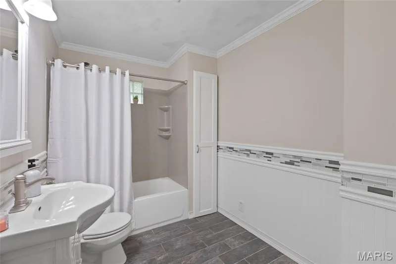Pedestal sink with a single-handle faucet, white bathtub with a built-in shower, wall-mounted storage shelves, and dark tile flooring — 712 Highland Drive, O'Fallon, MO 63366