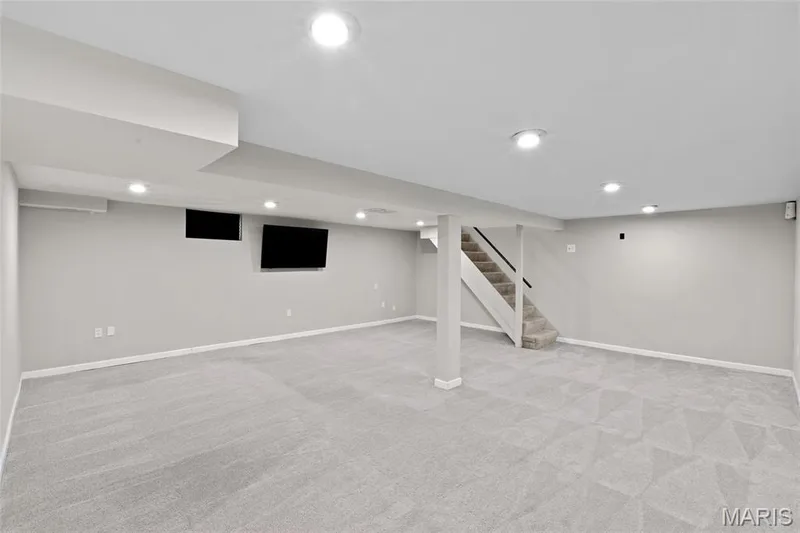 Recessed lighting, light gray wall paint, light gray patterned carpeting, staircase with dark handrail, and built-in wall-mounted television — 712 Highland Drive, O'Fallon, MO 63366