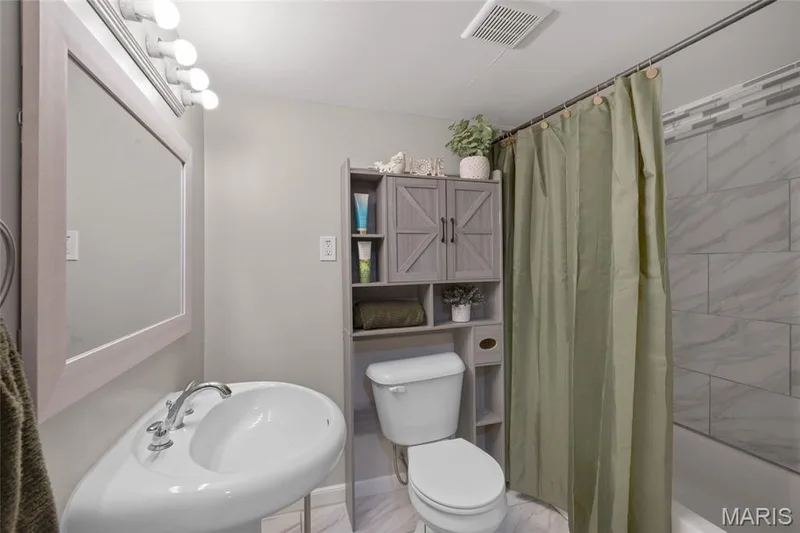 Pedestal sink with chrome faucet, framed vanity mirror, overhead vanity lighting, toilet with built-in storage cabinet, and tiled shower surround — 712 Highland Drive, O'Fallon, MO 63366