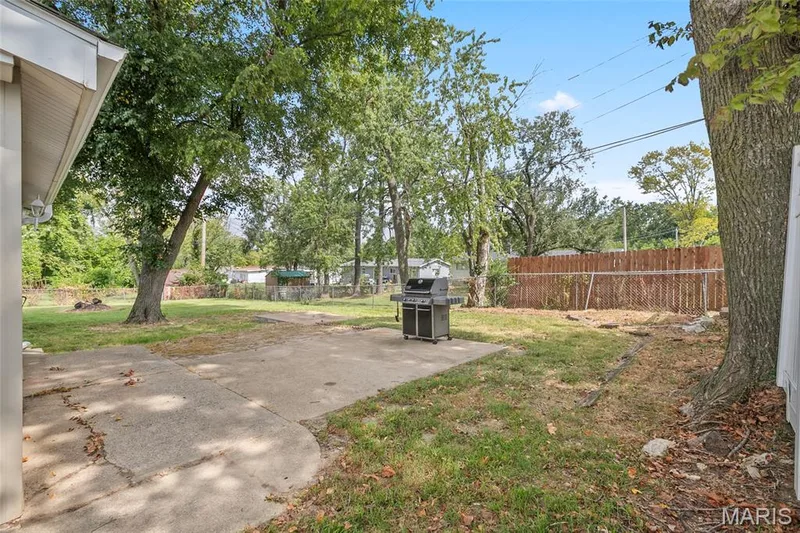 Expansive backyard featuring mature trees, a concrete patio, and a wood privacy fence — 712 Highland Drive, O'Fallon, MO 63366