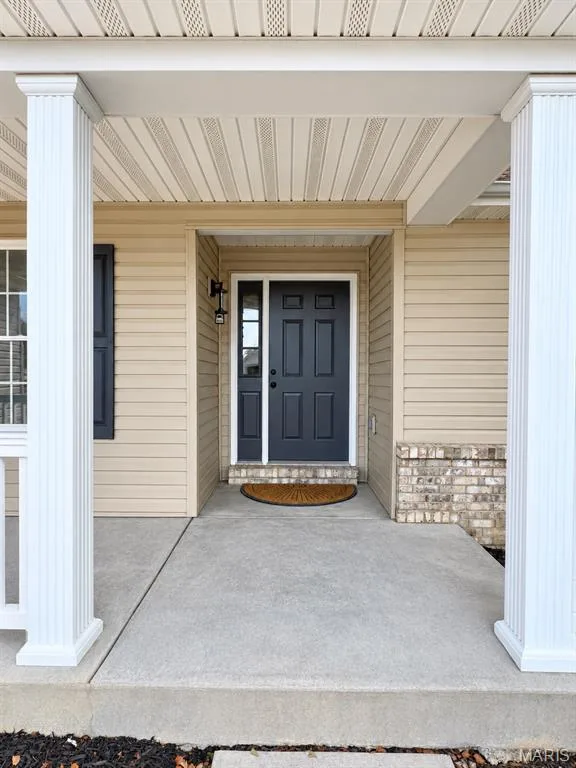 Covered front porch with white columns, light beige siding, brick facade, dark paneled entry door, and concrete porch — 1731 Fairview Farms Circle, Wentzville, MO 63385